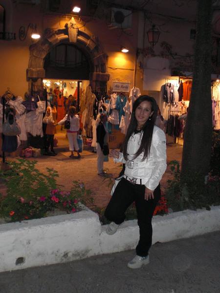 Woman in white jacket by a flower bed in front of lit clothing stalls