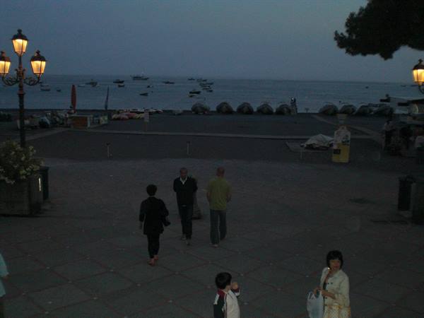 Seaside square at dusk, people walking, boats offshore, lit street lamps