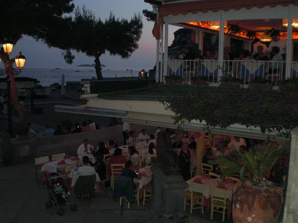 Busy seaside restaurant at dusk, set tables, warm lights, diners on terraces