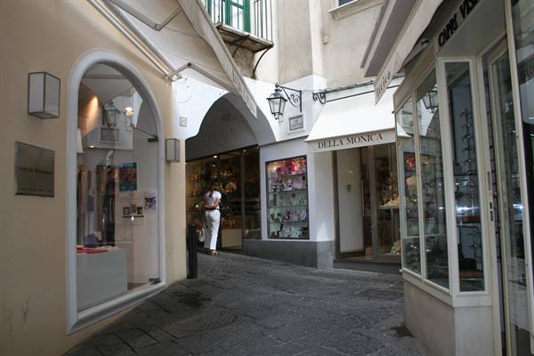 Woman walks uphill past boutiques, Della Monica sign, cobbled lane