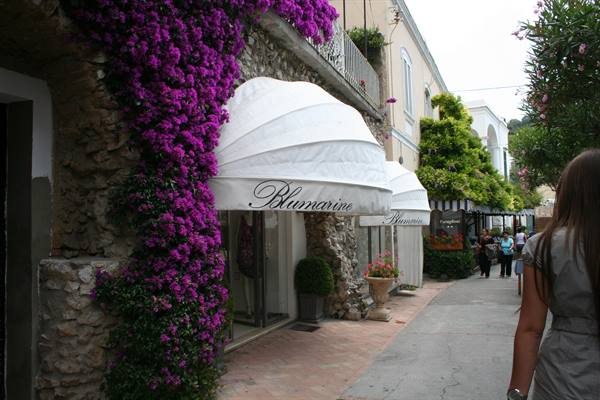 Alley with Blumarine boutique, white canopies and purple bougainvillea on stone wall, Capri
