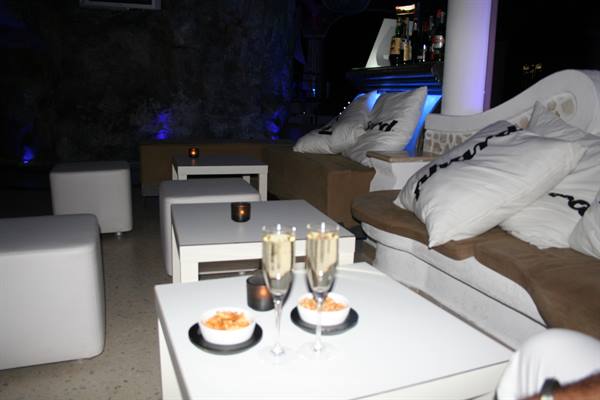 Two champagne glasses and snacks on white tables in a lounge with sofas and candles