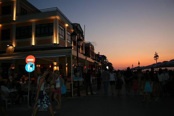 Crowded seaside promenade at dusk, lit buildings, people walking toward the water