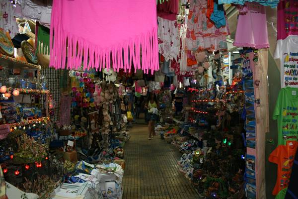 Crowded souvenir market aisle, pink fringed shawl hanging, colorful lights on stalls