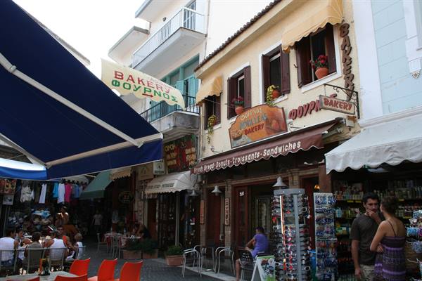 Greek bakery signs on yellow buildings, awnings and umbrellas over busy café tables