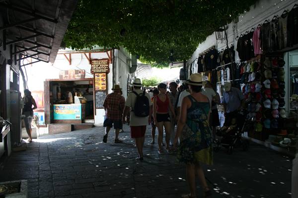 Tourists walking under shaded pergola along cobbled street lined with hat and t-shirt stalls