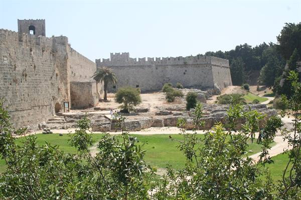 Medieval stone walls of Rhodes town, palm trees and green bushes under sun