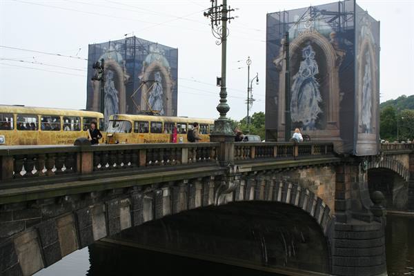 Yellow tram crossing the stone bridge, banner panels wrap the bridge piers