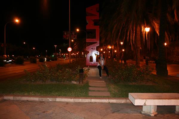 Girl sitting under the red Palma sign at night among palms and streetlights
