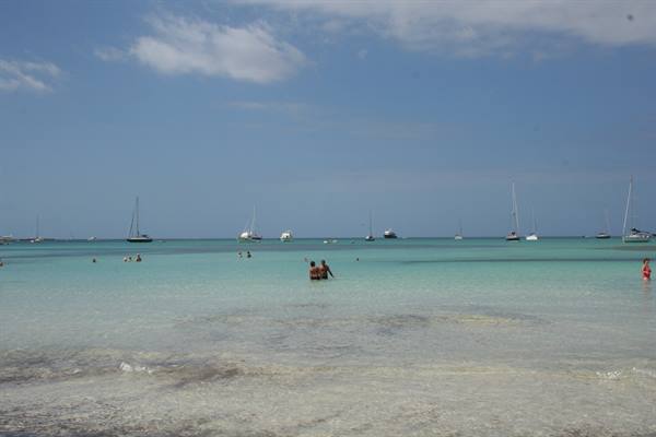 Swimmers in turquoise water, sailboats anchored on the horizon, pale sand foreground