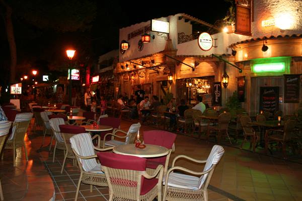 Empty rattan tables in front of illuminated night bars, people seated at tables