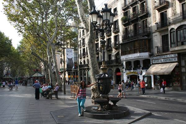 Woman beside Canaletes fountain on Barcelona's Rambla, trees and strollers around