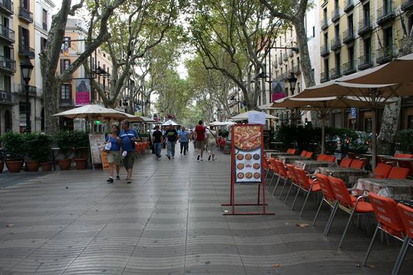 Tourists stroll under plane trees along Barcelona's Ramblas, with parasols and orange chairs