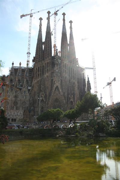 Sagrada Família in Barcelona, stone spires with cranes among trees and a pond