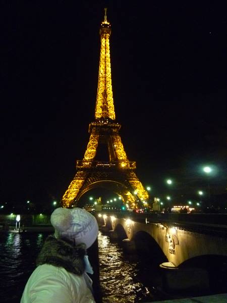 Woman in hat watches the illuminated Eiffel Tower from the Seine bridge