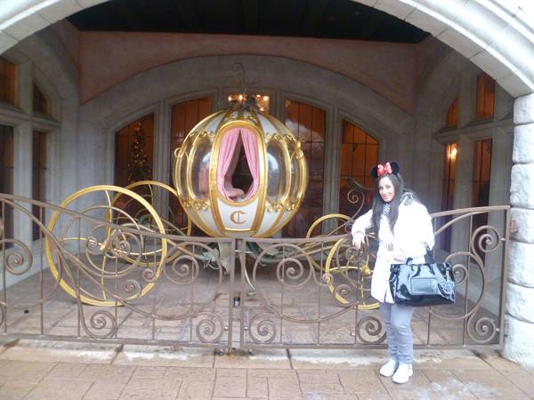 Woman beside Cinderella's golden pumpkin carriage under a stone archway