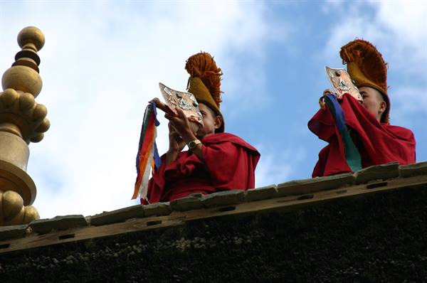 Monks in red robes blow long copper horns on monastery rooftop