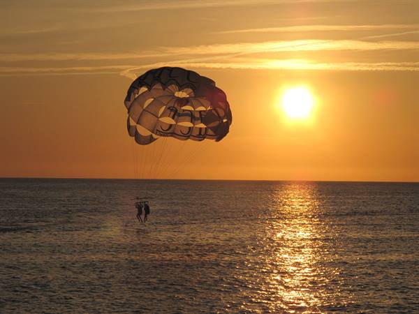 Couple parasailing with white canopy over golden sea, low sun glowing