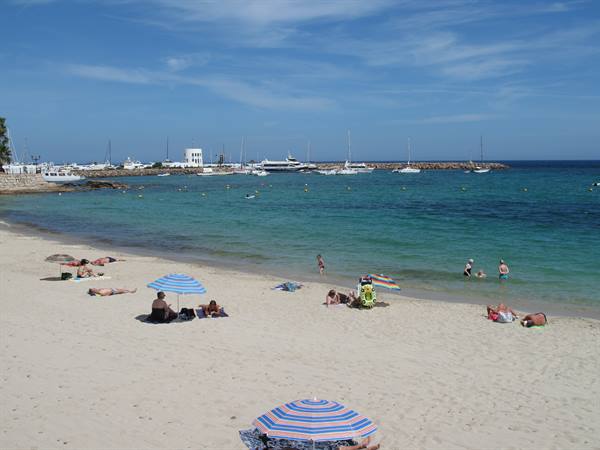 Beachgoers under striped umbrellas on pale sand, yachts beyond the breakwater