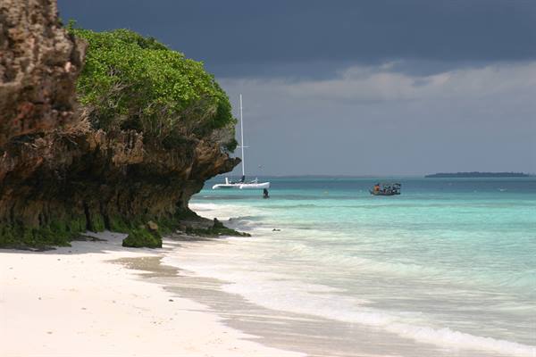 Vegetation-covered rocky cliff on white sand beach, catamaran and boats on turquoise water