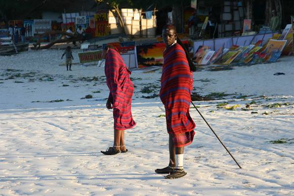 Two Maasai men wrapped in red shukas walk along the white sand beach
