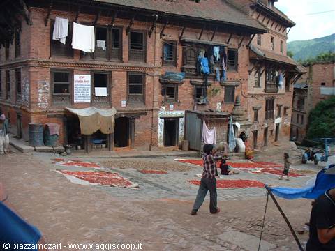 People cross a square with grain drying in the sun before brick houses