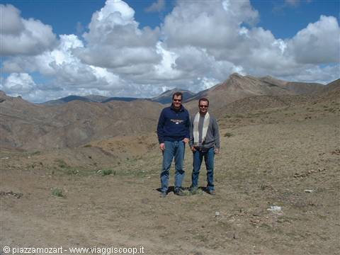 Two men in jeans stand on an arid plateau among mountains under white clouds