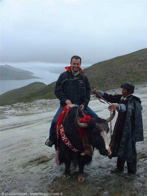 Tourist riding a yak with red saddle, local guide holding rope, foggy lake behind