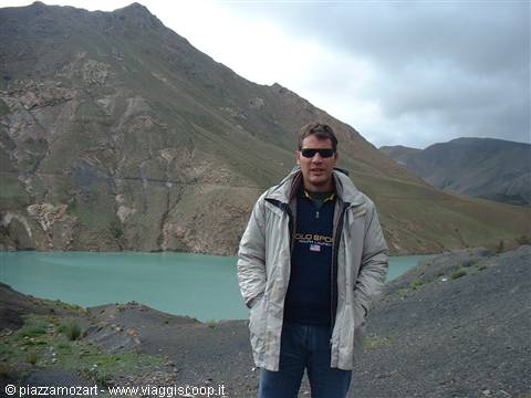 Man in jacket and sunglasses in front of a turquoise lake among barren mountains and cloudy sky