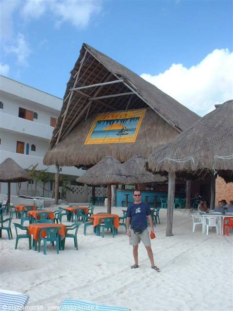 Man standing on sand in front of thatched palapa bar with orange tables