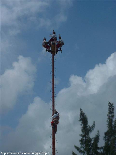 Voladores de Papantla atop the tall pole, one descending on rope, cloudy sky