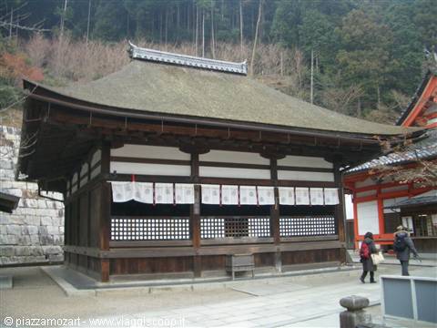 Wooden temple with thatched roof, white banners, visitors walking, forested hillside behind