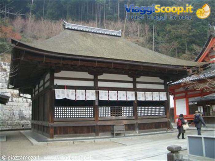 Wooden temple with thatched roof, white banners, visitors walking, forested hillside behind