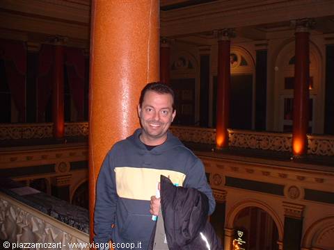 Smiling man near red column inside an ornate theatre with balconies