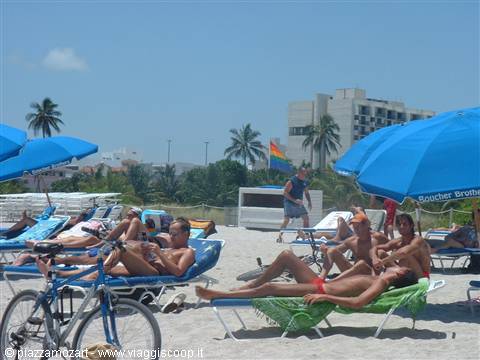 Sunbathers under blue umbrellas on white sand, palms and buildings