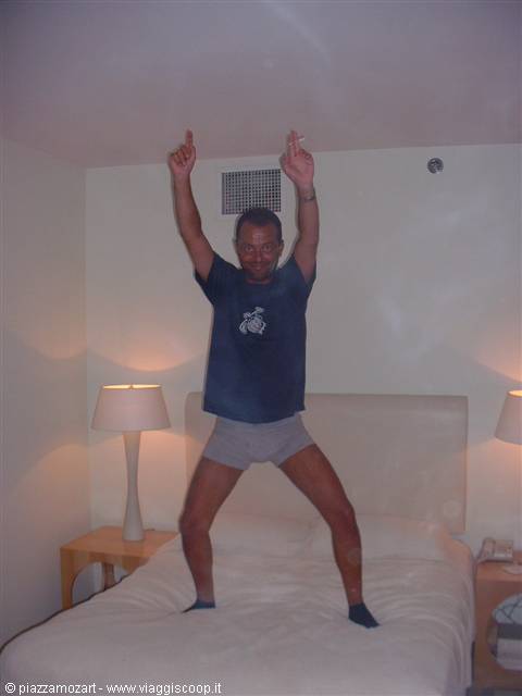 Man in t-shirt and boxers stands on hotel bed with arms raised