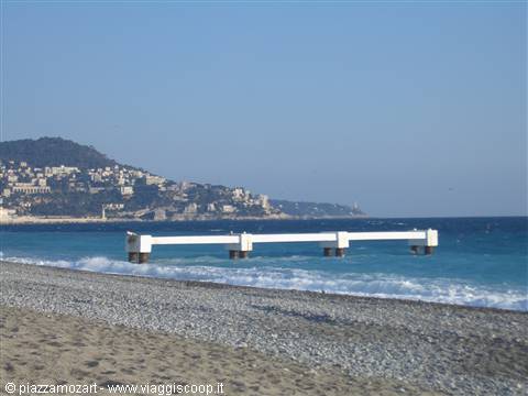 White pier over turquoise sea in Nice, pebble beach, hillside houses behind