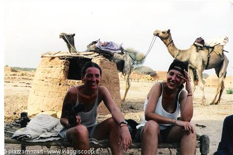 Two smiling women near mud huts and camels in the desert