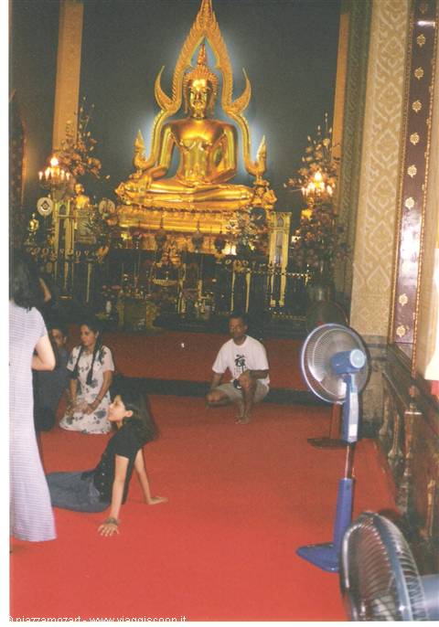 Tourists sitting on red carpet in front of a large golden Buddha in the temple