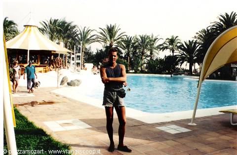 Young man in tank top beside the pool, palm trees and yellow tents
