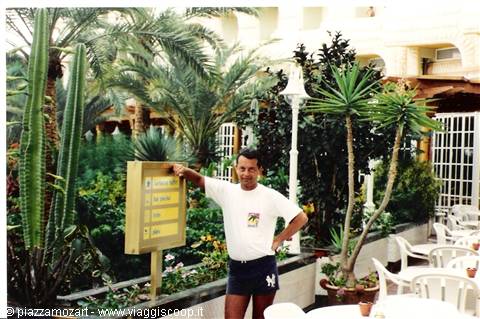 Man in white t-shirt beside wooden sign among palm trees and cacti