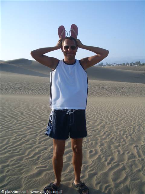 Man with sunglasses holds two flip-flops above his head among sand dunes