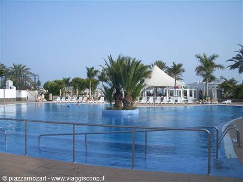 Blue pool with central palm island, white sun loungers and canopy tent under clear sky