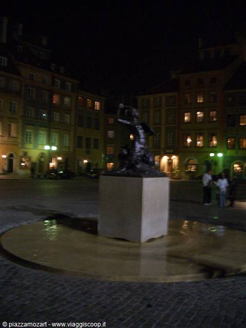 The Warsaw Mermaid statue at night, lit up in the cobbled square
