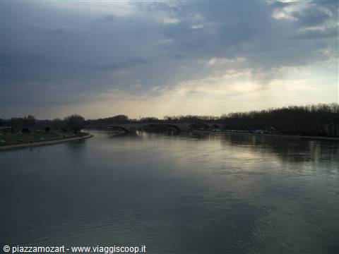 The Po river flows calm, stone arch bridge among bare trees, cloudy sky