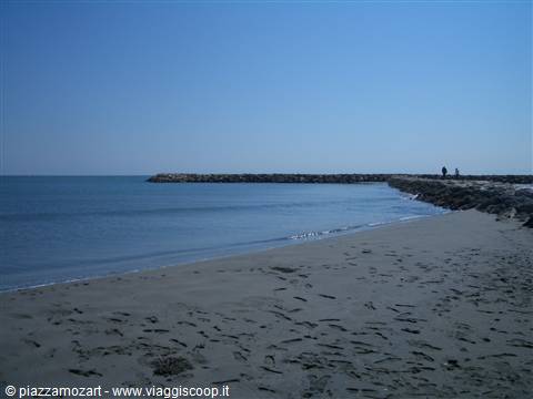 Dark sand beach with long rocky breakwater and two people walking on it