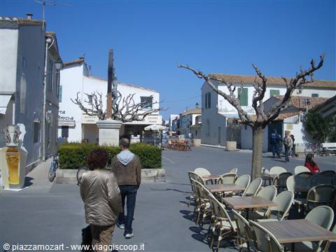 Square with empty bistro chairs and white houses, two people walking in the sun