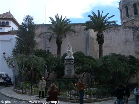 Marble statue among palm trees and old walls, tourists on the square