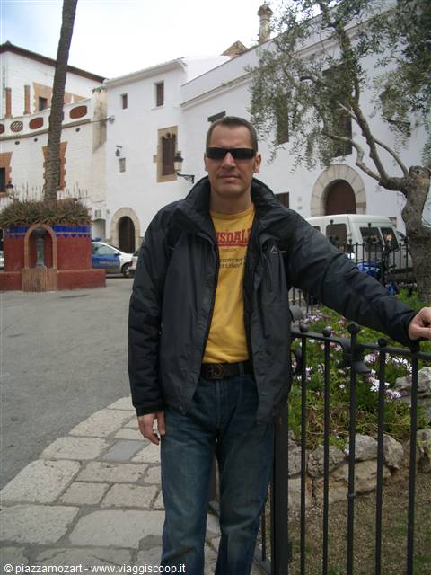 Man in jacket and sunglasses leaning on iron fence near whitewashed buildings