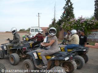 Helmeted riders on black quad bikes along a dusty dirt road near a village